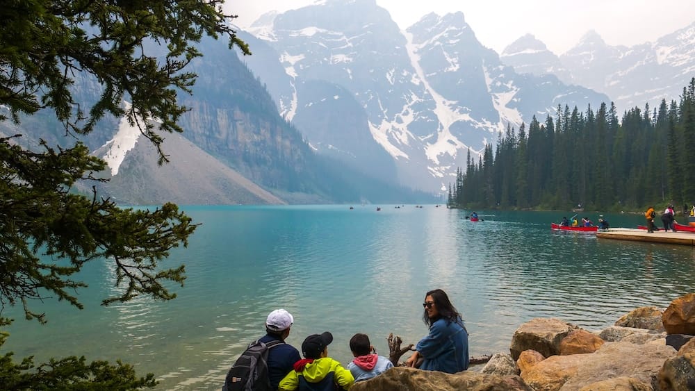Lake Louise shoreline in Banff National Park, Alberta, Canada with visitors, canoes and mountain scenery Visitors sit and stand along the rocky shore of Lake Louise in Banff National Park, Alberta, Canada, with red canoes on turquoise water and steep snow-streaked mountain peaks beyond.