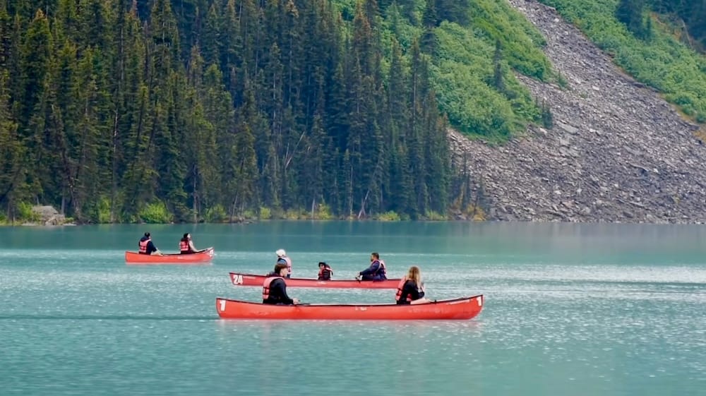 Red canoes paddling across turquoise Lake Louise in Banff National Park, Alberta, Canada, with forested shoreline and rocky mountain slopes rising behind the water.