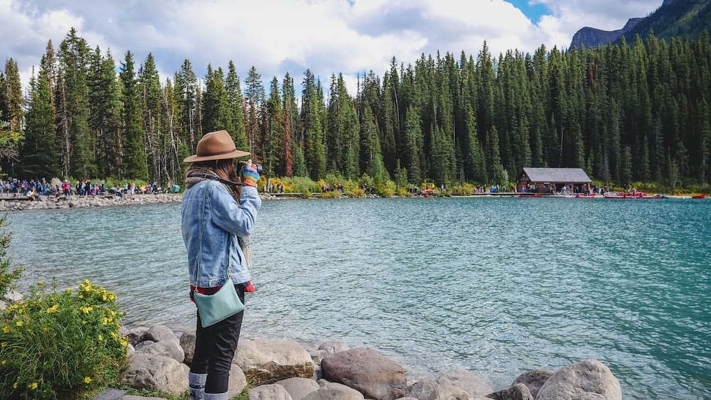 Audrey Bergner photographing Lake Louise in Banff National Park Alberta Canada from the turquoise lakeshore Audrey Bergner photographs Lake Louise in Banff National Park, Alberta, Canada, from the rocky shoreline, with turquoise water, evergreen forest, visitors and the lakeside boathouse visible across the lake.