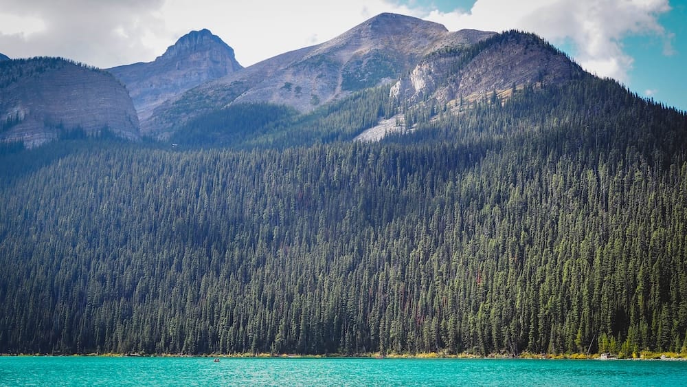 Turquoise Lake Louise in Banff National Park, Alberta, Canada, beneath dense evergreen forest and layered rocky mountain peaks under broken cloud and blue sky.