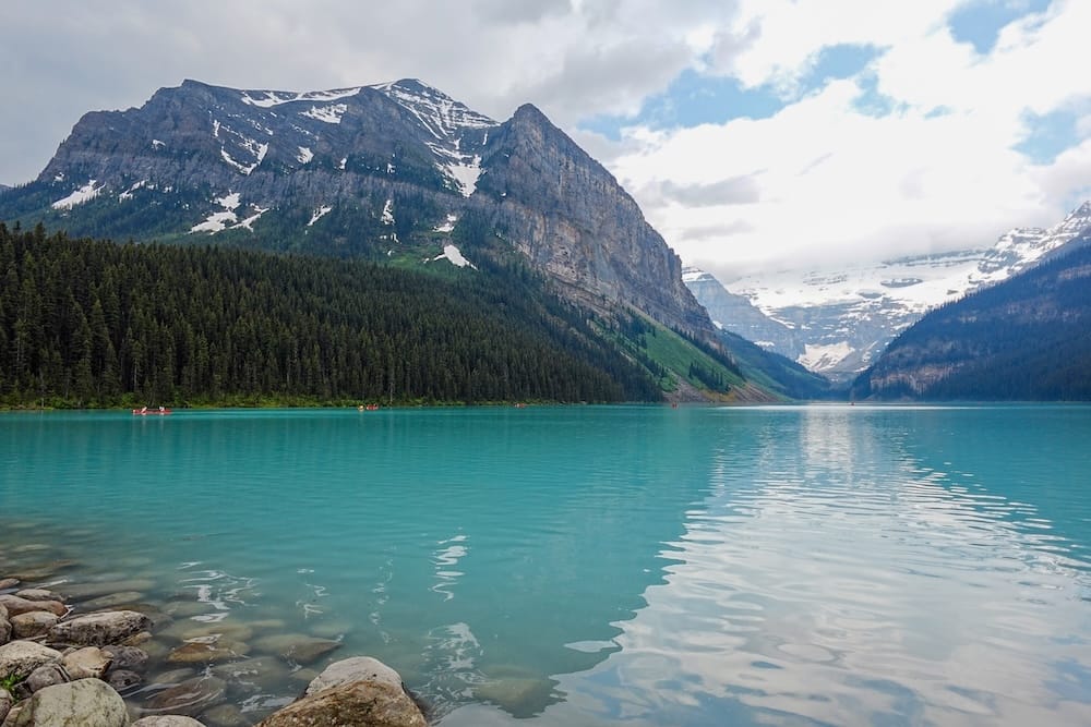 Turquoise Lake Louise in Banff National Park, Alberta, Canada, with a rocky shoreline, dense evergreen forest, steep mountain walls and a glacier visible beneath broken cloud in the distance.