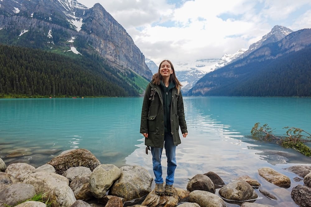 Audrey Bergner standing on rocks beside turquoise Lake Louise in Banff National Park, Alberta, Canada, with forested slopes, steep Rocky Mountain walls and snow-covered peaks behind her.