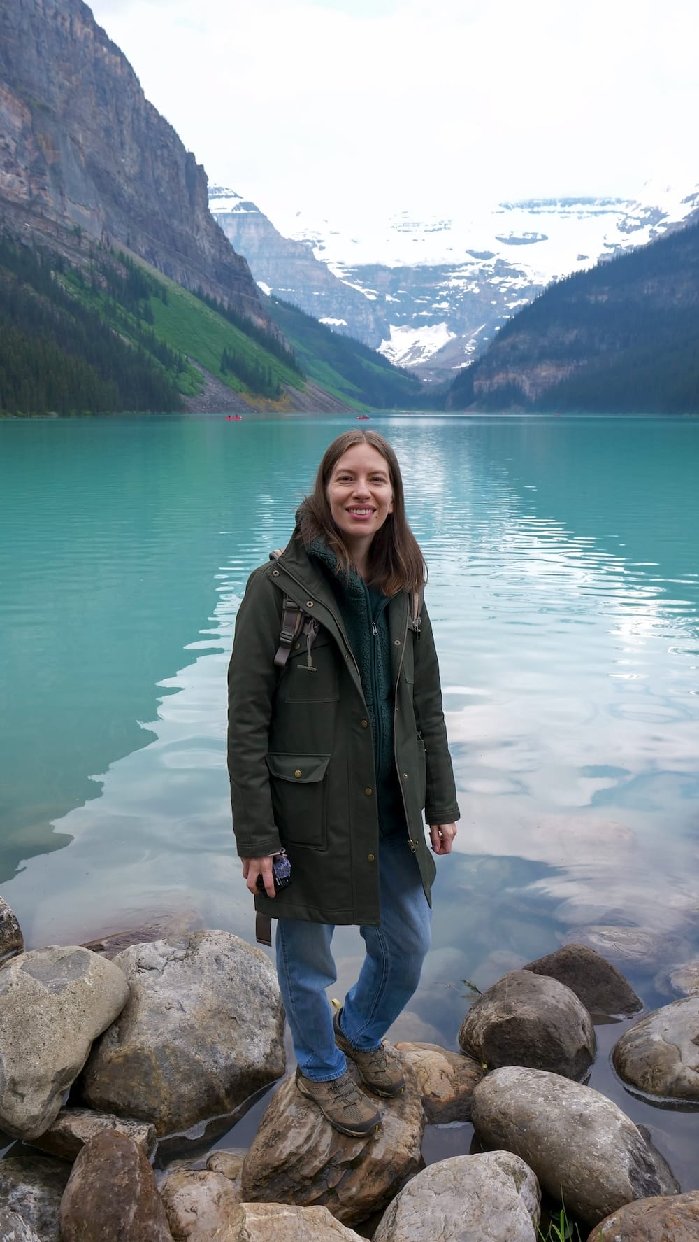 Audrey Bergner standing on rocks beside turquoise Lake Louise in Banff National Park, Alberta, Canada, with forested slopes, steep Rocky Mountain walls and snow-covered peaks behind her.