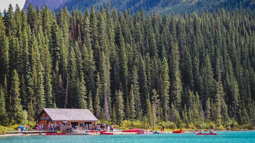 Lake Louise boathouse and canoe shoreline in Banff National Park, Alberta, Canada, with red canoes, visitors gathered at the dock, turquoise water in the foreground and a dense evergreen forest rising behind.