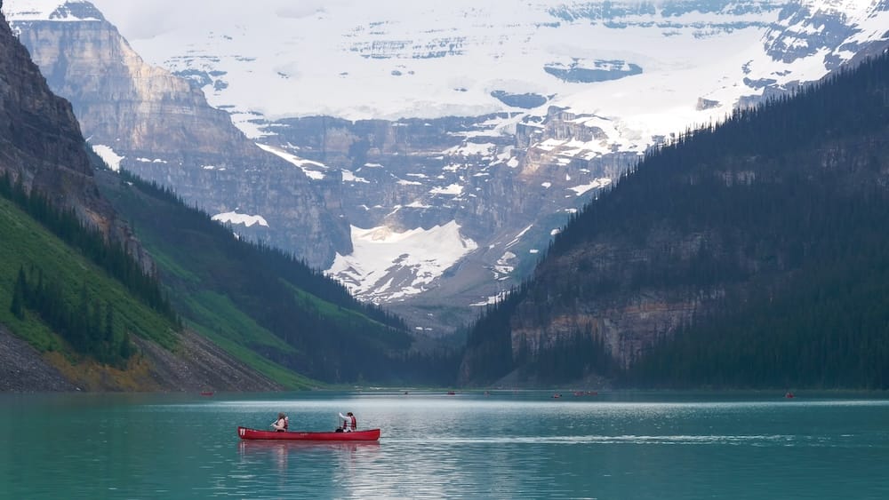 Canoeists paddle across the turquoise waters of Lake Louise in Banff National Park, Alberta, Canada, framed by glacier-capped mountains rising above the shoreline.