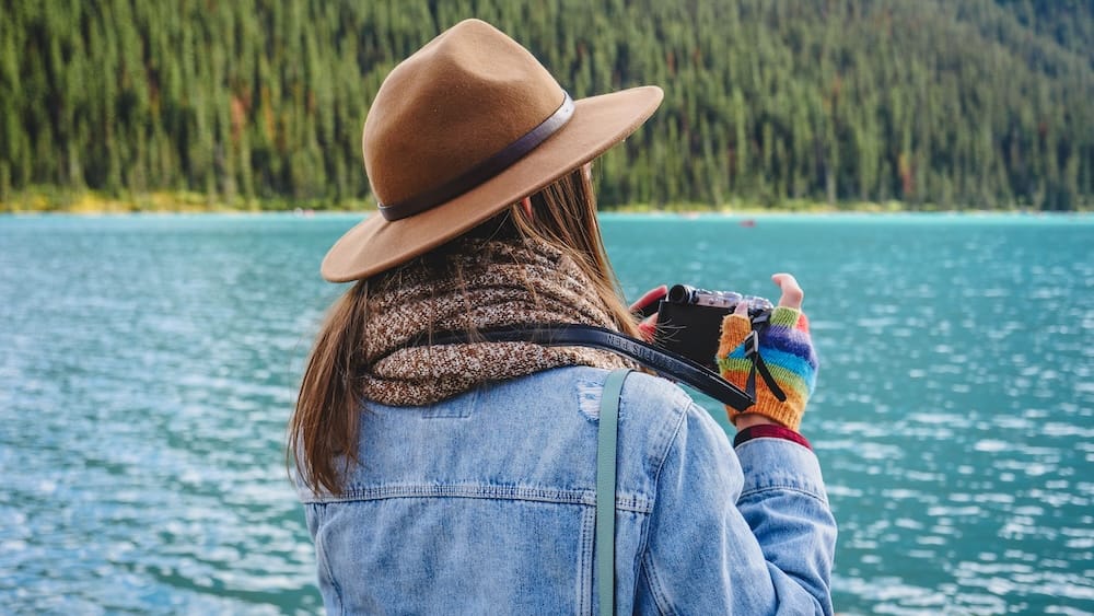 Audrey Bergner photographing turquoise Lake Louise from the Lakeshore Trail in Banff National Park, Alberta, Canada, wearing a brown hat and denim jacket with forested slopes across the water.