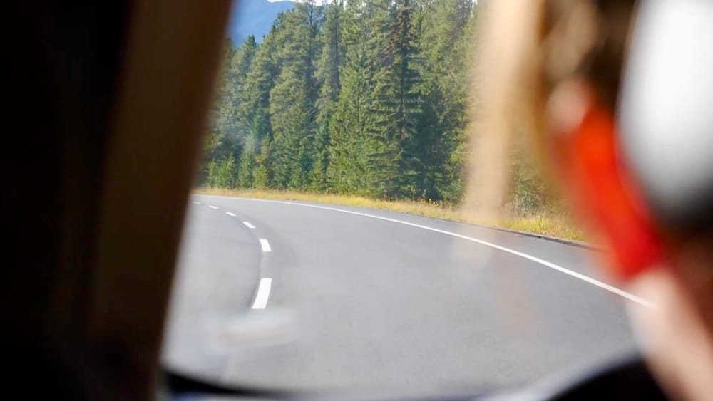 View through a vehicle window along a curving mountain road near Lake Louise in Banff National Park, Alberta, Canada, with evergreen forest lining the roadside and a passenger blurred in the foreground.