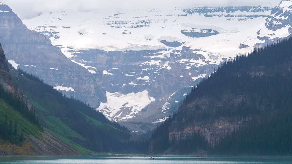 Victoria Glacier rising above the far end of Lake Louise in Banff National Park, Alberta, Canada, with snow-covered icefields, steep mountain walls and small red canoes visible on the turquoise water below.