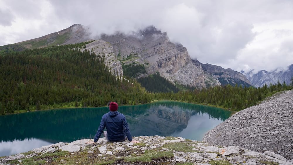 Samuel Jeffery at Lake Matthias in Banff National Park, Alberta, Canada on a backcountry hike from Halfway Lodge Samuel Jeffery sits above Lake Matthias in Banff National Park, Alberta, Canada, looking across dark turquoise water toward forested slopes and steep rocky mountains wrapped in low cloud.