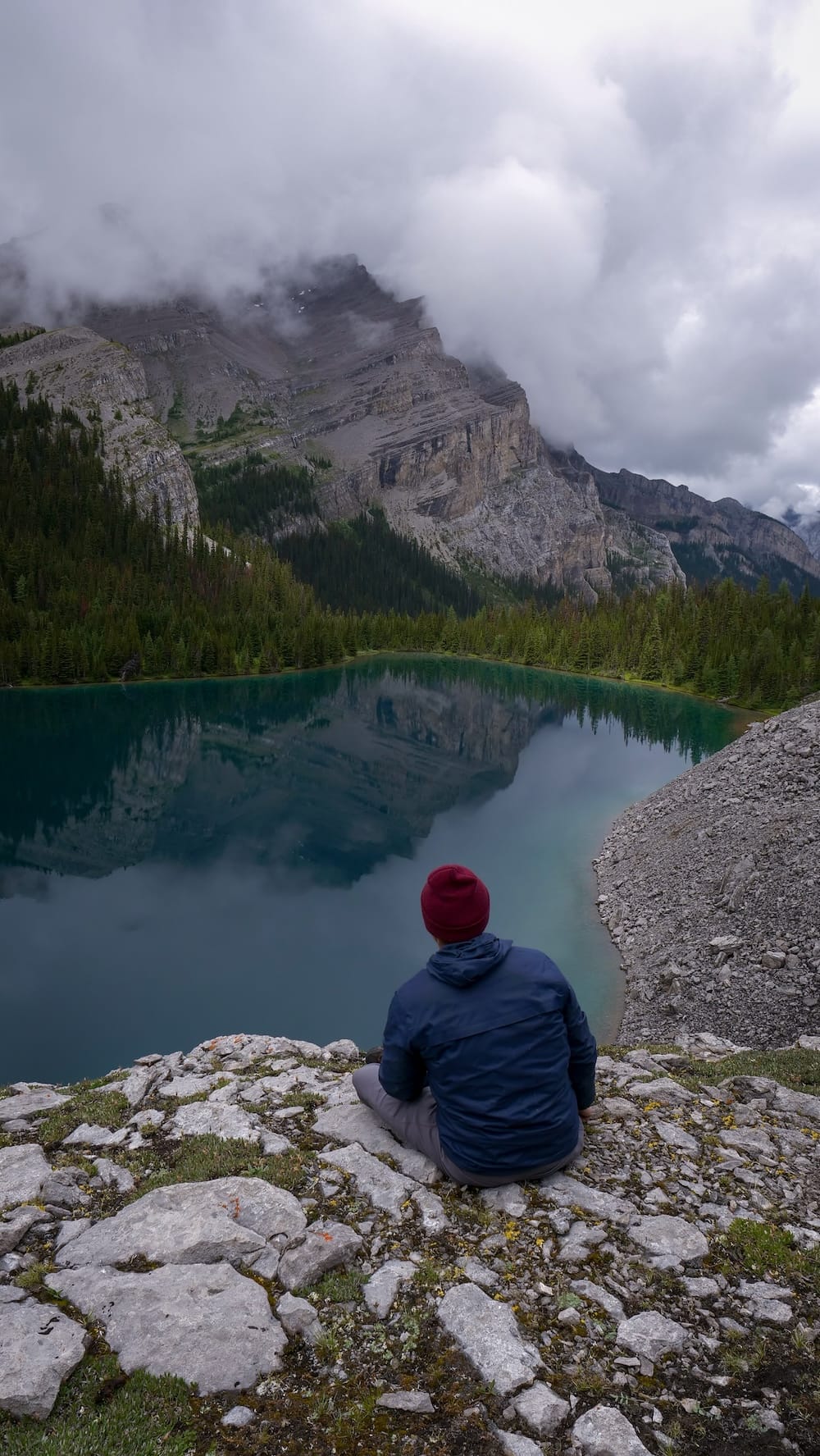 Samuel Jeffery overlooking Lake Matthias in Banff National Park, Alberta, Canada during the six-day backcountry trip Samuel Jeffery sits on a rocky overlook above Lake Matthias in Banff National Park, Alberta, Canada, facing a dark turquoise alpine lake beneath steep mountain walls partly wrapped in low cloud.