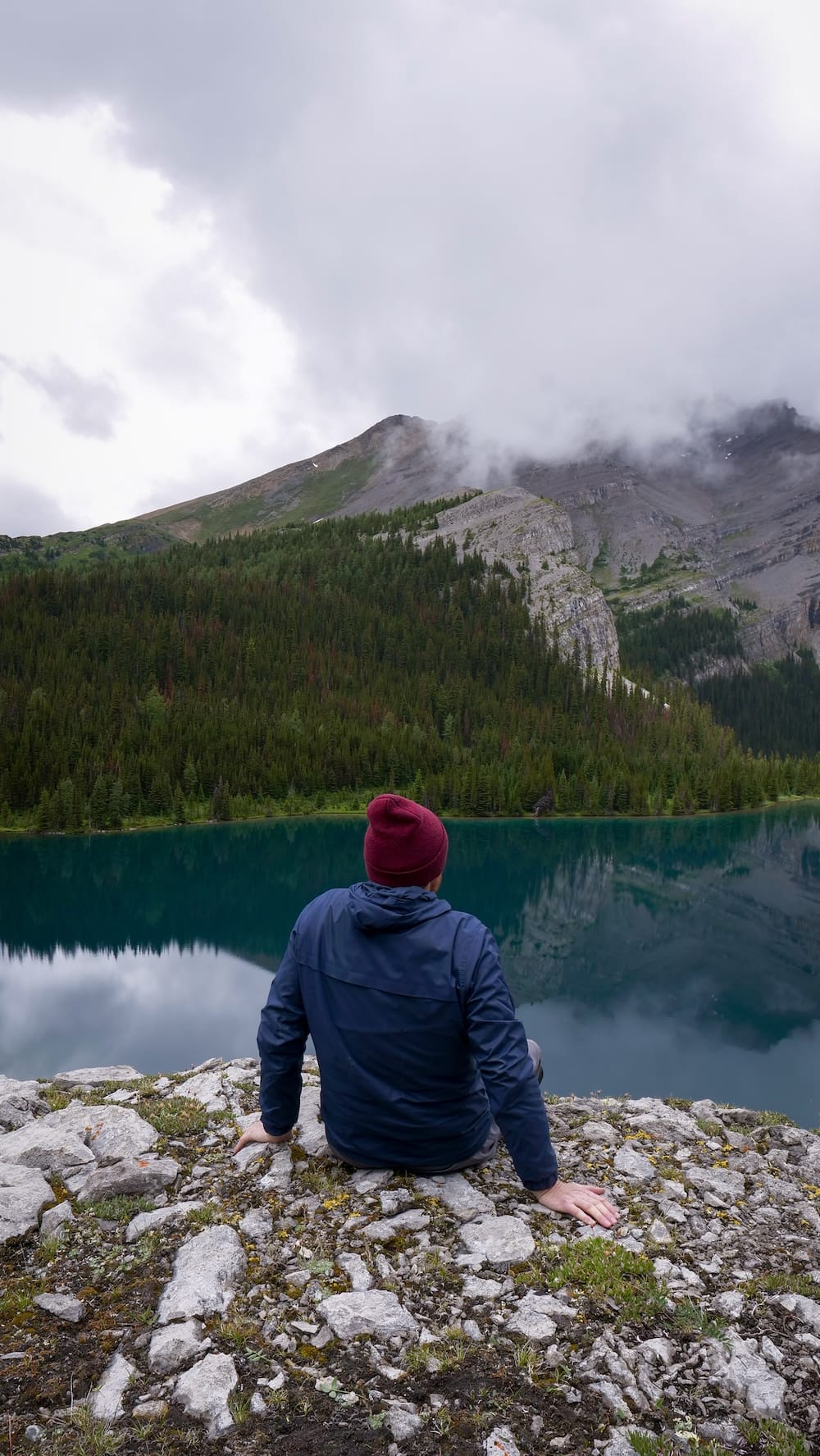 Samuel Jeffery está sentado sobre una saliente rocosa sobre Lake Matthias, en el Parque Nacional Banff, Alberta, Canadá, mirando el agua turquesa oscura, laderas boscosas y picos cubiertos por nubes.