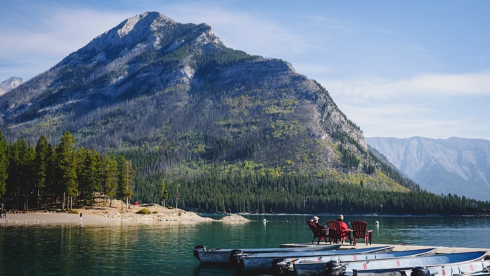 Rental boats line a dock at Lake Minnewanka in Banff National Park, Alberta, Canada, with calm blue-green water, a forested shoreline and steep Rocky Mountain slopes rising behind the lake.