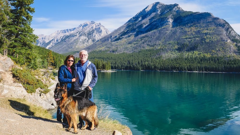 Maria and Daniel Bergner stand with Togo beside Lake Minnewanka in Banff National Park, Alberta, Canada, with clear blue-green water, forested shoreline and rugged mountain slopes behind them.