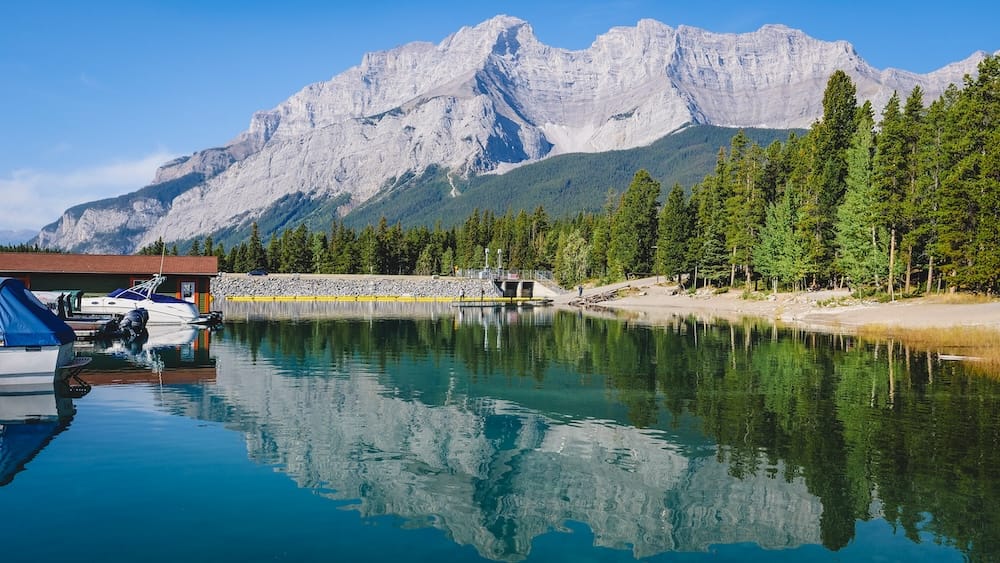 Boats and marina facilities sit along Lake Minnewanka in Banff National Park, Alberta, Canada, with calm blue water reflecting evergreen forest and a massive rocky mountain backdrop.