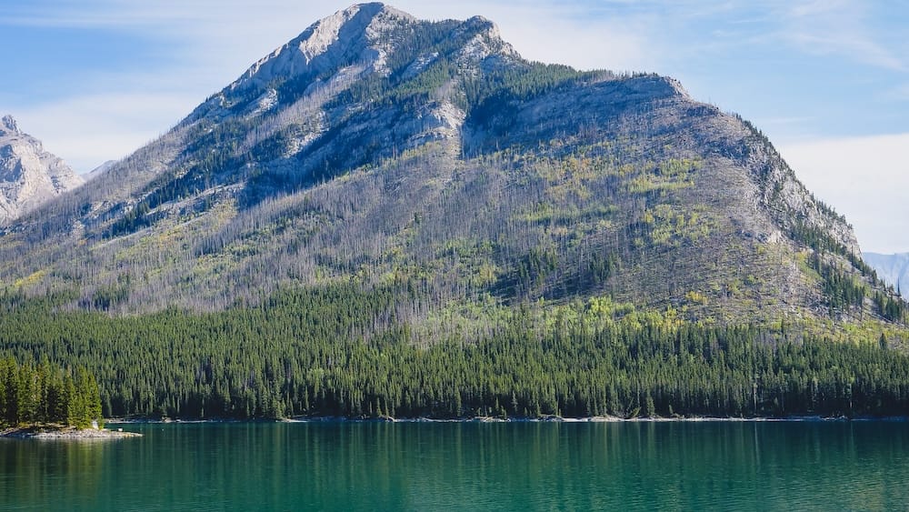 Lake Minnewanka in Banff National Park, Alberta, Canada with mountain slopes, forest and calm blue-green water Lake Minnewanka in Banff National Park, Alberta, Canada, with calm dark-turquoise water below a broad forested mountain slope and evergreen shoreline under a clear sky.