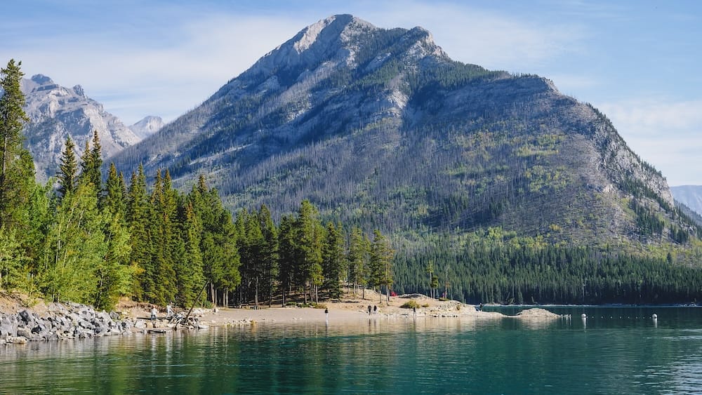 Lake Minnewanka in Banff National Park, Alberta, Canada with mountain lake, evergreen forest and shoreline scenery Lake Minnewanka in Banff National Park, Alberta, Canada, with calm blue-green water, an evergreen shoreline and a broad forested Rocky Mountain rising behind the lake under clear skies.