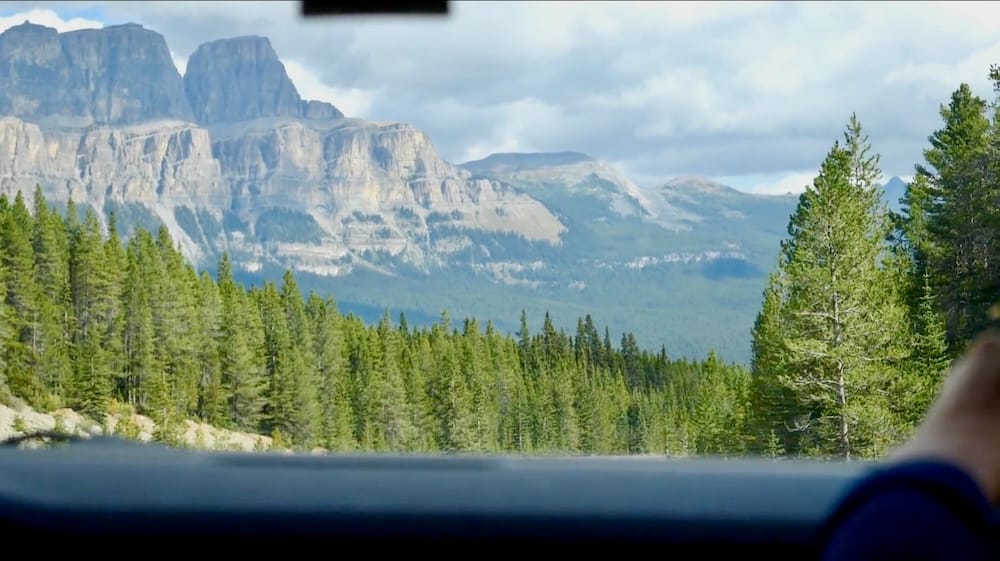 Mountain and forest scenery seen through a vehicle window while driving toward Lake Minnewanka in Banff National Park, Alberta, Canada, with layered rocky peaks rising above dense evergreen forest.