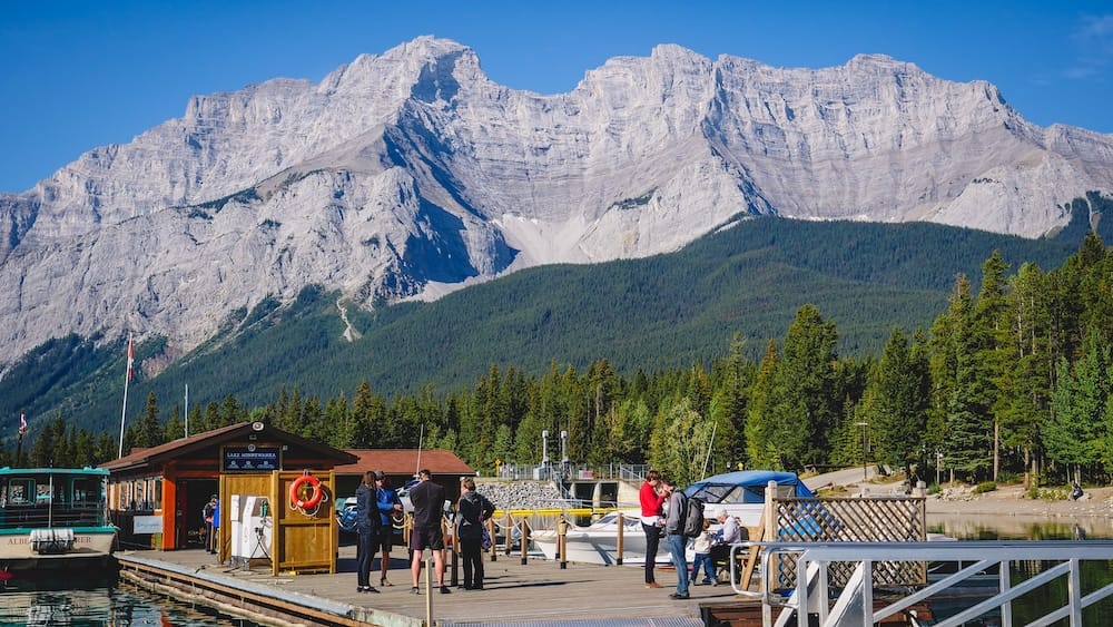 Muelle de botes del Lake Minnewanka, Parque Nacional Banff, Alberta, Canadá El muelle de botes del Lake Minnewanka en Alberta, Canadá, con visitantes y embarcaciones cerca del edificio de alquiler y una gran montaña que se eleva justo detrás del lago.