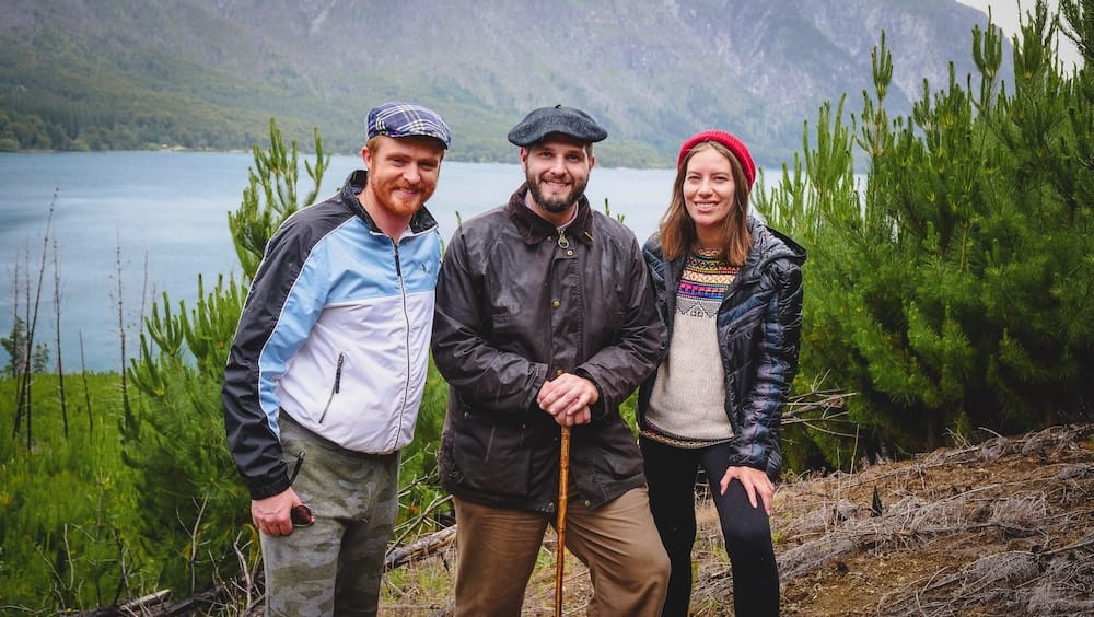 Samuel Jeffery and Audrey Bergner with a Local Friend Above Lake Puelo, El Bolsón, Río Negro, Patagonia Samuel Jeffery, Audrey Bergner, and a local friend stand smiling above turquoise Lake Puelo near El Bolsón, Río Negro, in Argentine Patagonia, with burnt tree stumps and young pines in front of forested mountains under cloudy skies.
