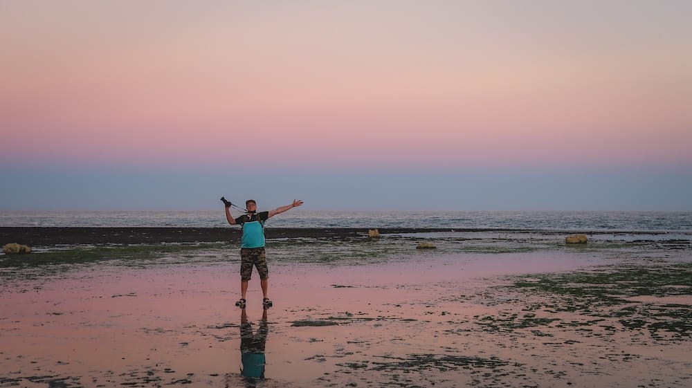 Las Grutas Patagonia sunset tidal flats with Samuel Jeffery enjoying peaceful Atlantic coastal reflections Las Grutas, Río Negro Patagonia — Samuel Jeffery stands on tidal flats at sunset with arms outstretched, reflecting pastel skies and calm Atlantic waters, capturing the surreal coastal beauty and wide-open feeling of this underrated beach destination.