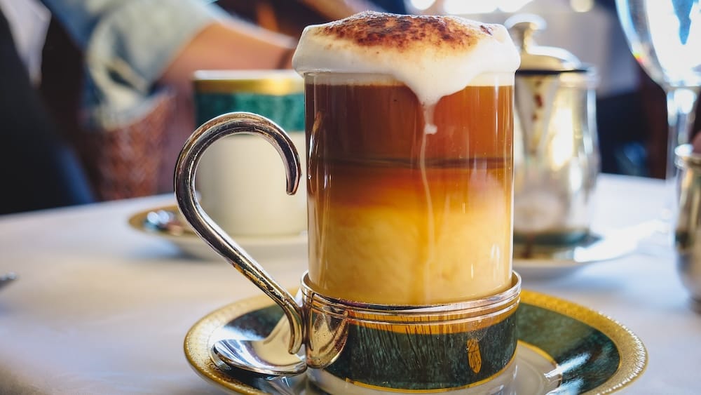 Layered Cappuccino Served During Afternoon Tea at Llao Llao Hotel, Bariloche, Río Negro, Argentine Patagonia A layered caramel cappuccino topped with cinnamon-dusted foam sits in a glass mug with a silver handle on an ornate gold-and-green saucer during afternoon tea at Llao Llao Hotel's Winter Garden in Bariloche, Río Negro, Argentine Patagonia, Argentina.