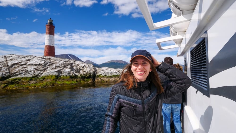Audrey Bergner stands on a Beagle Channel cruise beside Les Éclaireurs Lighthouse near Ushuaia, Tierra del Fuego, Patagonia, Argentina, with rocky islets, dark coastal water and mountains behind her.