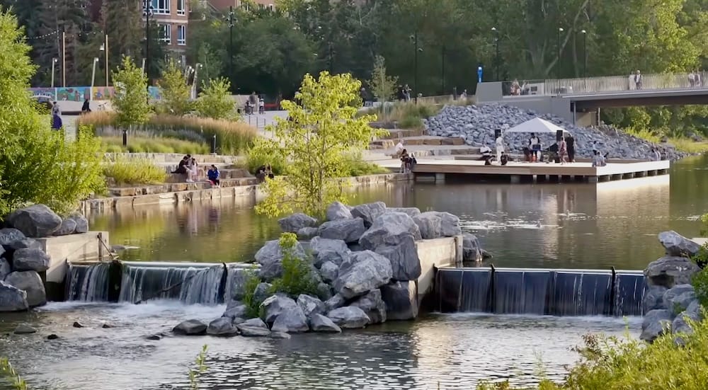 Música en vivo junto al dique, Prince's Island Park, Calgary, Alberta, Canadá Gente reunida en un pequeño escenario junto al agua y en escalones de piedra para ver música en vivo junto a un dique en el río Bow en Prince's Island Park, Calgary, Alberta, Canadá, con un puente peatonal y árboles verdes de fondo.