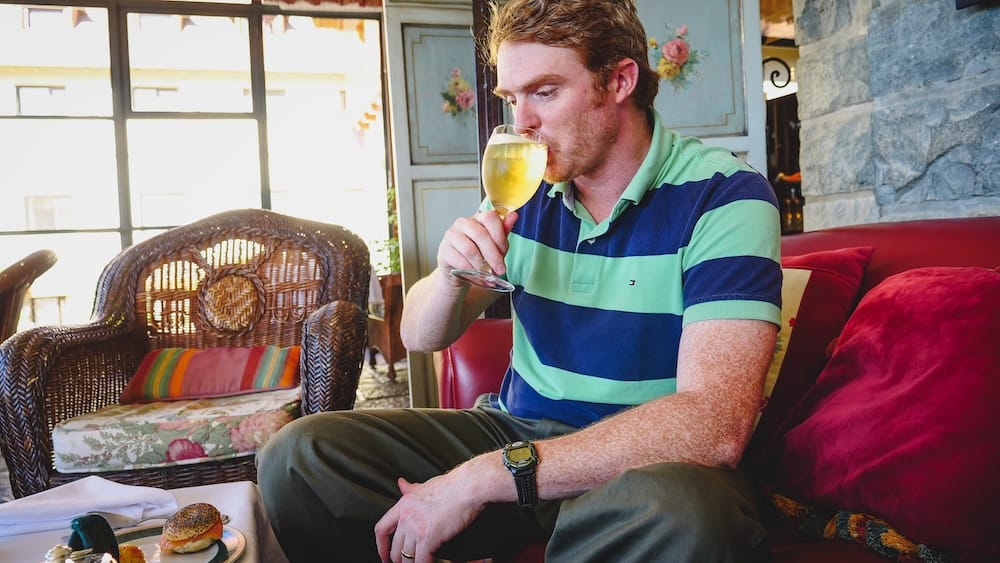 Samuel Jeffery sips a glass of white wine inside Llao Llao Hotel in Bariloche, Río Negro, Argentina, seated beside wicker furniture, stone walls, floral panels and warm alpine-style decor.