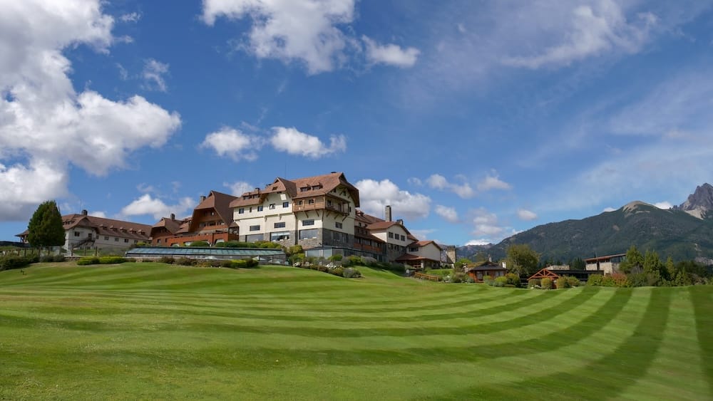 Llao Llao Hotel rises above striped green lawns in Bariloche, Río Negro, Patagonia, Argentina, with alpine-inspired timber-and-stone architecture, landscaped gardens and mountain peaks beneath a bright blue sky.