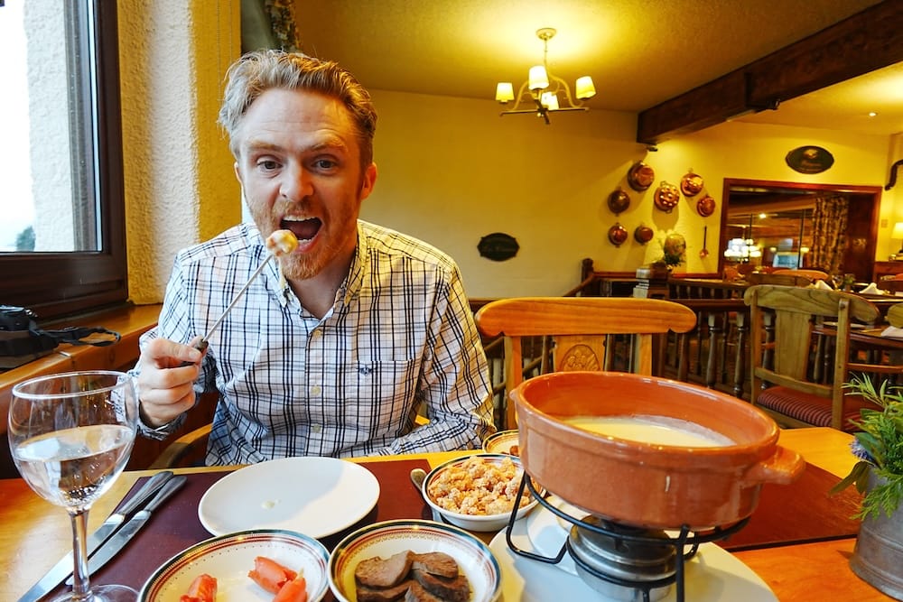 Samuel Jeffery eating cheese fondue at Llao Llao Hotel in Bariloche, Río Negro, Argentina, seated at a wooden table with the fondue pot, side dishes and a warm alpine-style dining room in the background.
