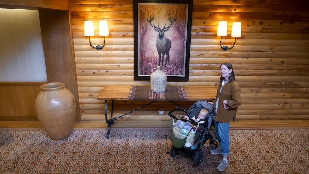 Audrey Bergner stands with toddler Aurelia Jeffery in a stroller inside Llao Llao Hotel in Bariloche, Río Negro, Argentina, beside timber walls, patterned carpets, pottery and alpine wildlife artwork.