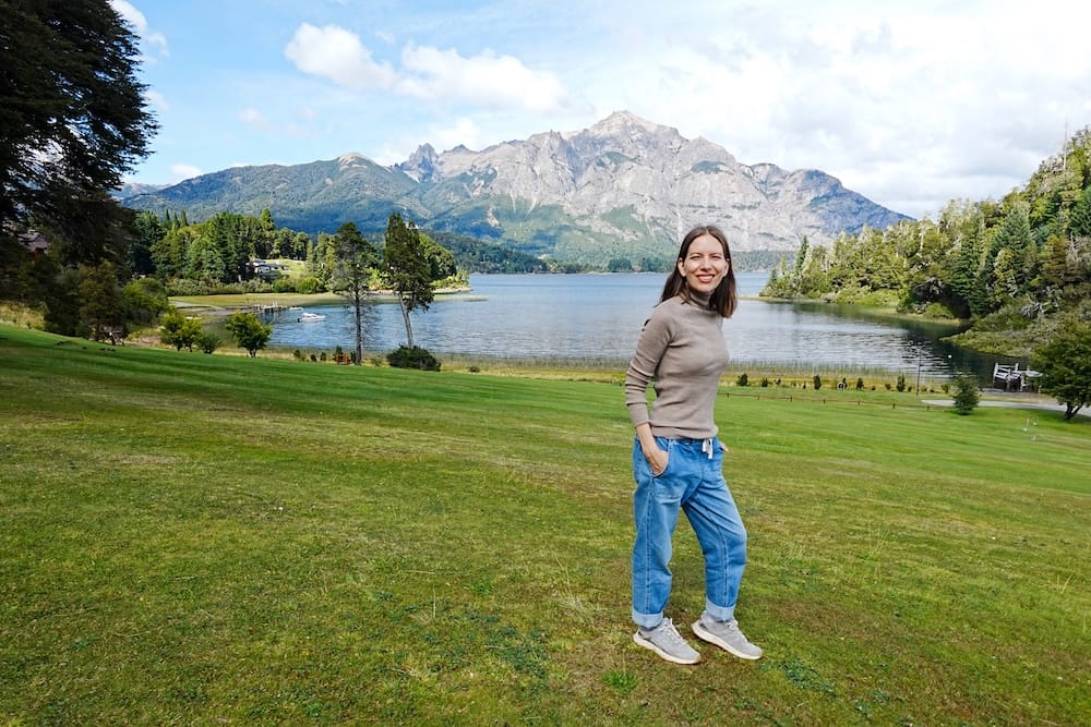 Audrey Bergner stands on the green grounds of Llao Llao Hotel in Bariloche, Río Negro, Argentina, with a calm lake, dense forest and dramatic Patagonian mountain peaks rising behind her.