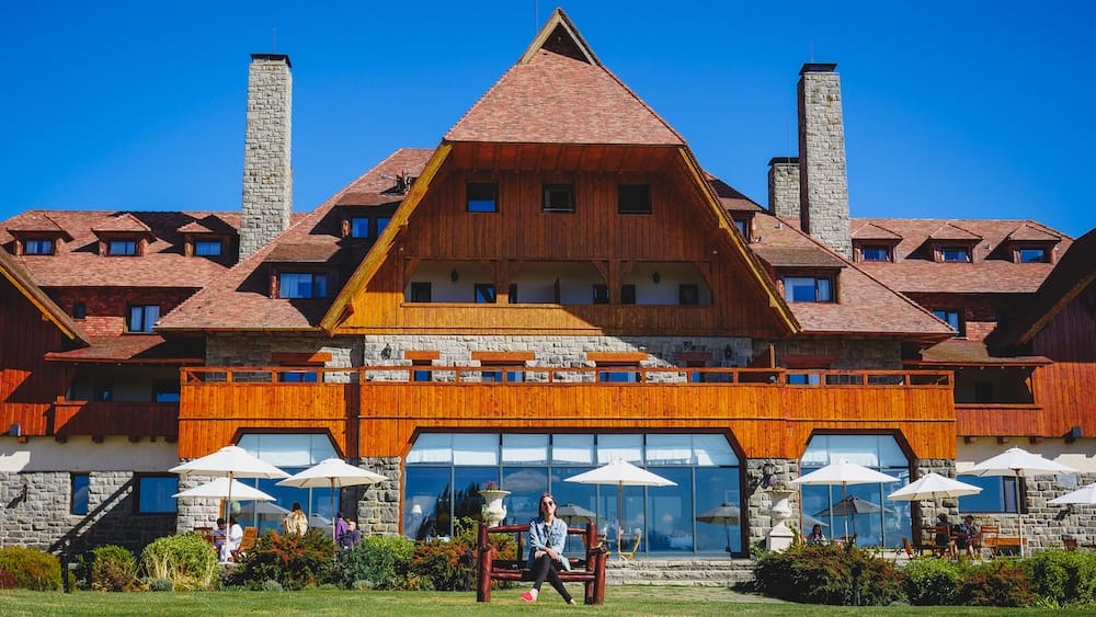 Audrey Bergner Outside Llao Llao Hotel in Bariloche, Río Negro, Patagonia, Argentina Audrey Bergner sits on a wooden bench outside Llao Llao Hotel in Bariloche, Río Negro, Patagonia, Argentina, with the resort’s steep alpine roofs, stone chimneys, timber balconies and terrace seating behind her.