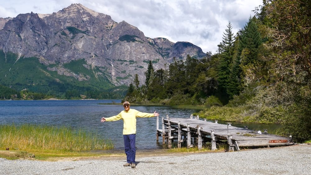 Samuel Jeffery stands with arms out beside a wooden lakeside pier on the grounds of Llao Llao Hotel in Bariloche, Río Negro, Argentina, with forest, reeds and a dramatic Patagonian mountain behind him.