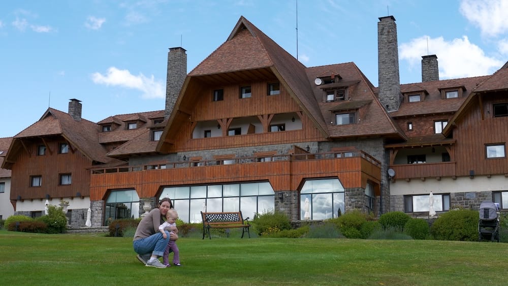 Bustillo Building Exterior, Llao Llao Hotel, Bariloche, Río Negro, Argentine Patagonia, with Audrey Bergner and Aurelia Jeffery Audrey Bergner crouches with toddler Aurelia Jeffery on the lawn outside the stone-and-wood Bustillo building at Llao Llao Hotel in Bariloche, Río Negro, Argentine Patagonia, under a partly cloudy sky.