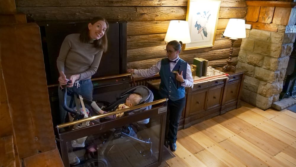 Audrey Bergner stands beside Aurelia Jeffery in a stroller inside a wood-panelled elevator at Llao Llao Hotel in Bariloche, Río Negro, Patagonia, Argentina, while a uniformed staff member operates the lift.
