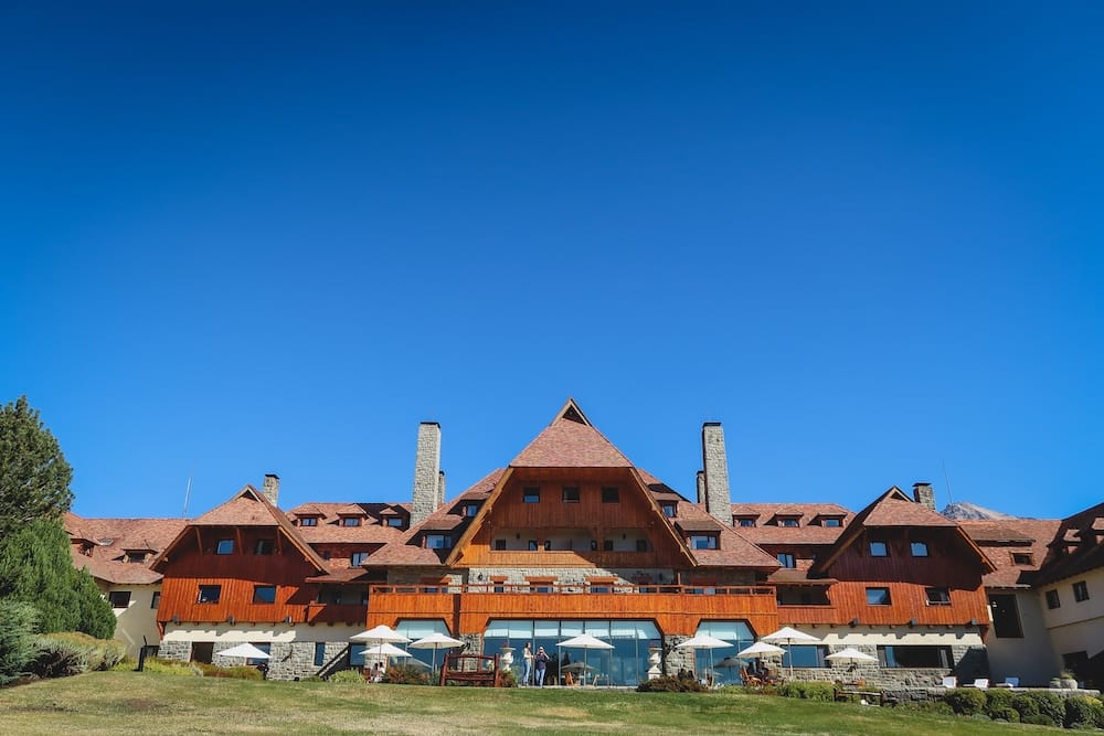 Llao Llao Hotel exterior, alpine wood-and-stone architecture, Bariloche, Río Negro, Argentine Patagonia, Argentina Llao Llao Hotel's steep-roofed alpine lodge exterior, built from dark wood and stone, with chimneys and an umbrella-shaded lawn terrace, set against a clear sky near Bariloche, Río Negro, in Argentine Patagonia.