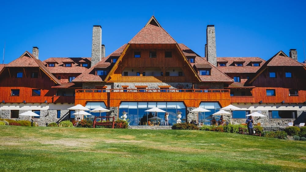 Llao Llao Hotel's Alpine Stone-and-Wood Facade and Front Lawn, Bariloche, Río Negro, Patagonia The steep-gabled, stone-and-wood exterior of Llao Llao Hotel rises behind a manicured lawn and umbrella-shaded terrace tables in Bariloche, Río Negro, Argentine Patagonia, Argentina, under a clear blue sky.