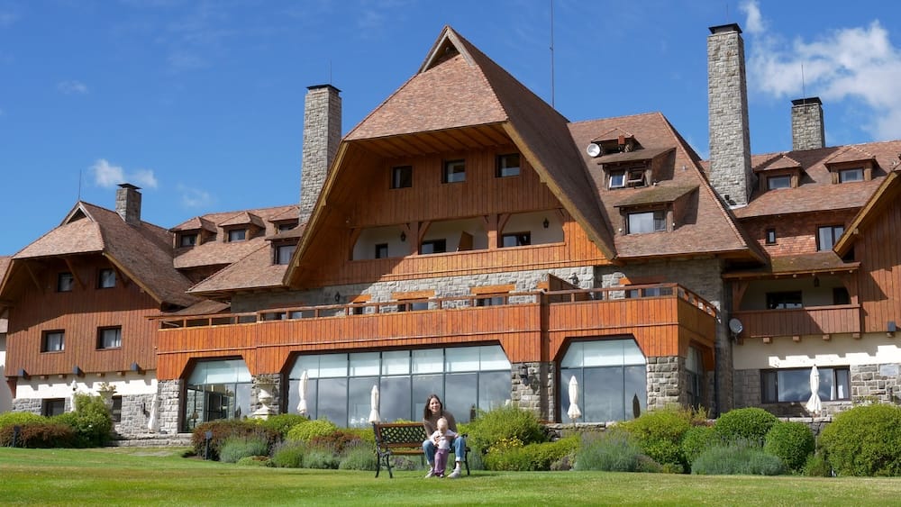 Audrey Bergner sits with daughter Aurelia Jeffery on a lawn bench in front of the Llao Llao Hotel exterior, showing its steep shingled roofs, stone base, and wood balconies, San Carlos de Bariloche, Argentine Patagonia.
