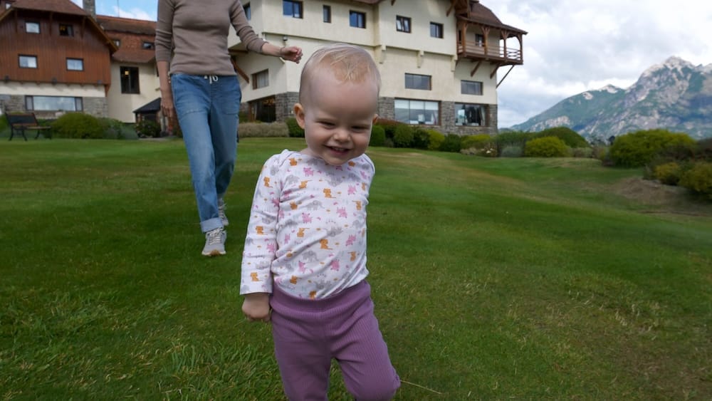 Aurelia Jeffery walks across the gardens at Llao Llao Hotel in Bariloche, Río Negro, Patagonia, Argentina, with Audrey Bergner following behind and the resort and mountain scenery visible in the background.