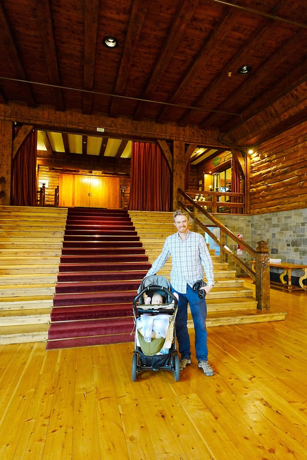 Samuel Jeffery stands beside Aurelia Jeffery in a stroller before a broad timber staircase inside Llao Llao Hotel in Bariloche, Río Negro, Patagonia, Argentina, surrounded by rustic log walls and polished wooden floors.