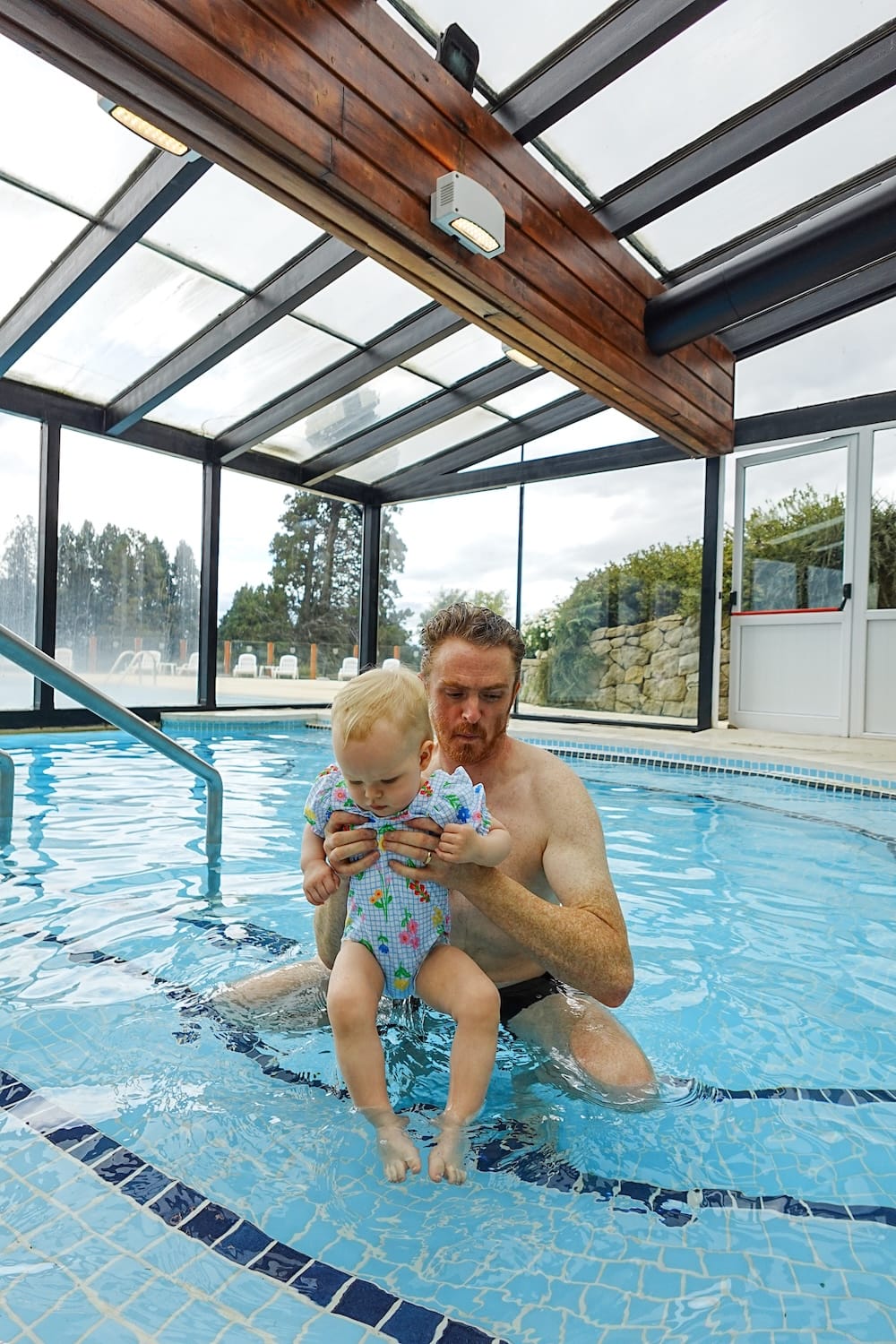 Samuel Jeffery holds Aurelia Jeffery in the heated indoor pool at Llao Llao Hotel in Bariloche, Río Negro, Patagonia, Argentina, with glass walls, pale blue water and the outdoor pool area visible behind them.
