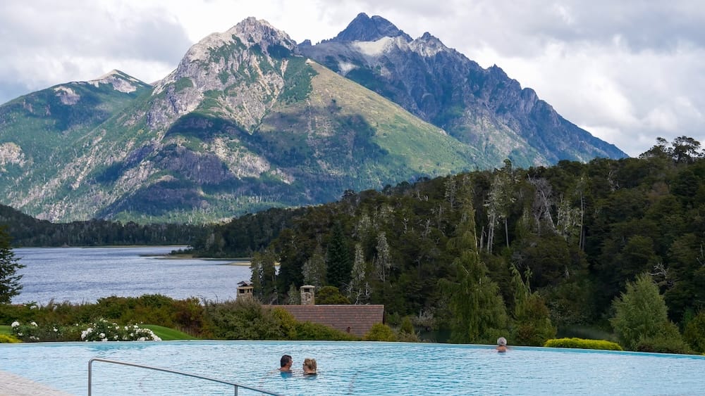 Guests swim in the outdoor infinity pool at Llao Llao Hotel in Bariloche, Río Negro, Patagonia, Argentina, with a lake, dense forest and steep Andean mountain peaks beyond the water.