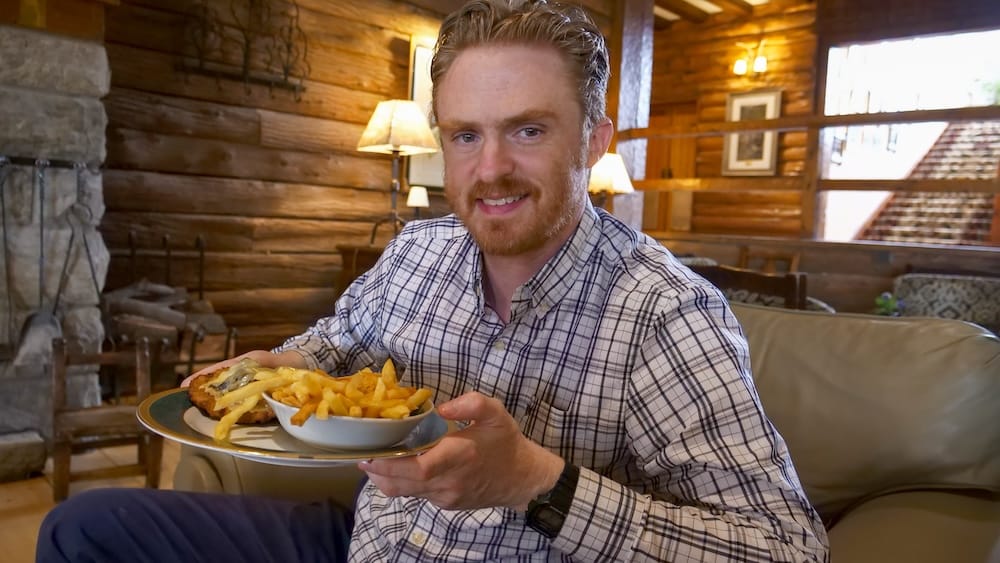 Samuel Jeffery Eating Milanesa and Fries at the Lobby Bar, Llao Llao Hotel, Bariloche, Río Negro, Patagonia Samuel Jeffery holds a plate of milanesa and fries while dining at the Lobby Bar inside Llao Llao Hotel in Bariloche, Río Negro, Patagonia, Argentina, with wood-panelled walls, warm lighting and alpine lodge décor around him.