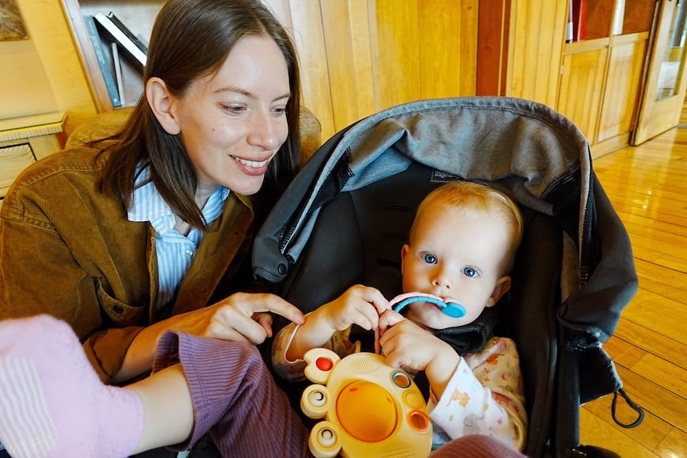 Audrey Bergner sits beside Aurelia Jeffery in a stroller inside Llao Llao Hotel in Bariloche, Río Negro, Patagonia, Argentina, as the toddler holds colourful toys against a backdrop of warm wooden cabinetry.