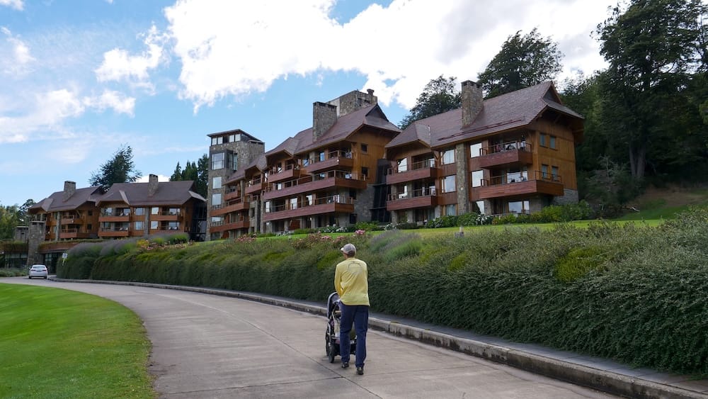 Samuel Jeffery pushes Aurelia Jeffery in a stroller beside the Moreno Wing at Llao Llao Hotel in Bariloche, Río Negro, Argentina, with timber balconies, stone walls and landscaped grounds.