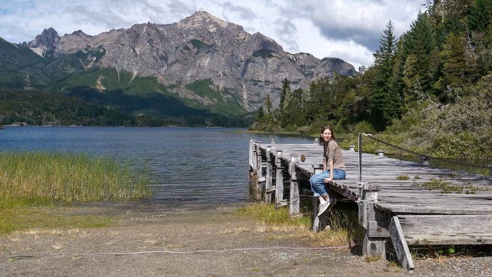 Audrey Bergner sits on a weathered wooden pier beside a mountain lake near Llao Llao Hotel in Bariloche, Río Negro, Patagonia, Argentina, with forest and dramatic alpine peaks behind her.