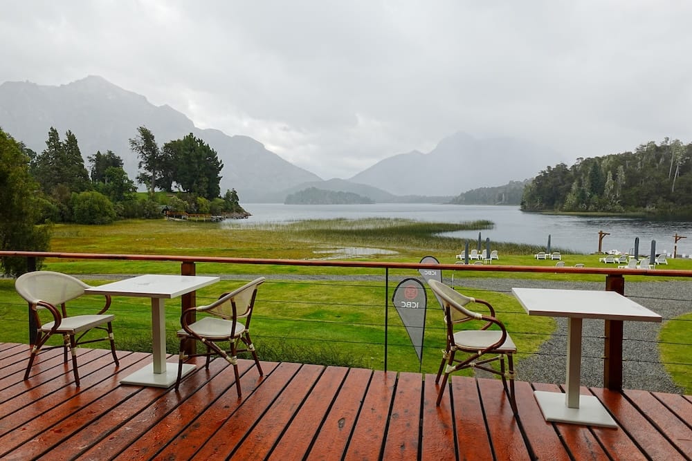 Wet wooden terrace tables overlook a misty lake, lawns and forested mountain slopes at Llao Llao Hotel in Bariloche, Río Negro, Patagonia, Argentina, beneath heavy rain clouds.