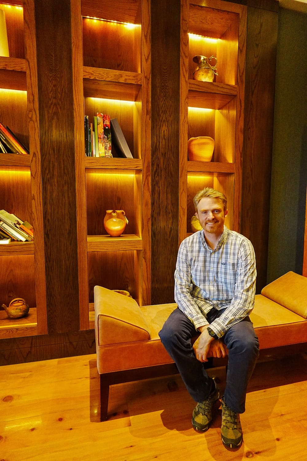 Samuel Jeffery sits on a cushioned bench beside illuminated wooden shelving, books and ceramic decor inside a refined lounge at Llao Llao Hotel in Bariloche, Río Negro, Patagonia, Argentina.