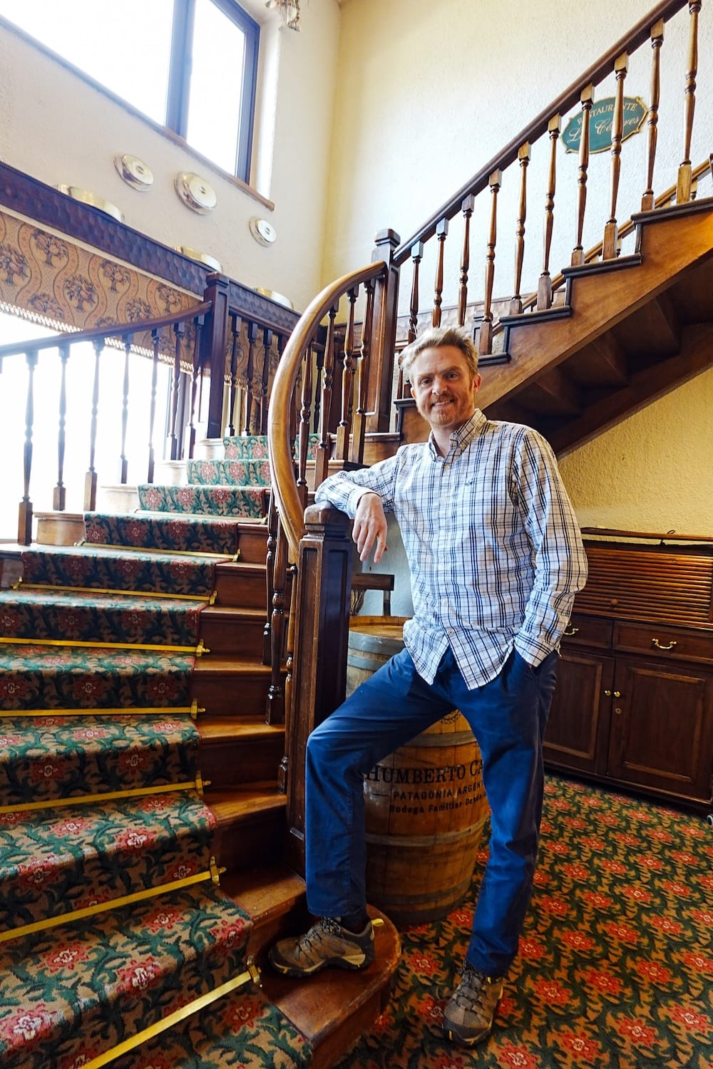 Samuel Jeffery standing beside a curved wooden staircase inside Llao Llao Hotel in San Carlos de Bariloche, Río Negro, Argentina, with patterned carpet, carved railings and a wine barrel.